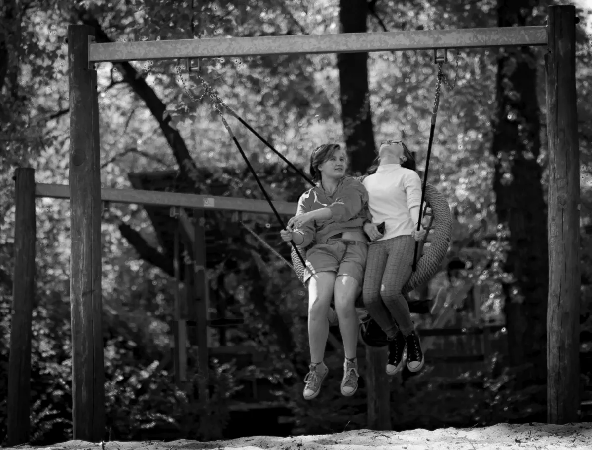 Two children sharing a round swing in a shaded playground